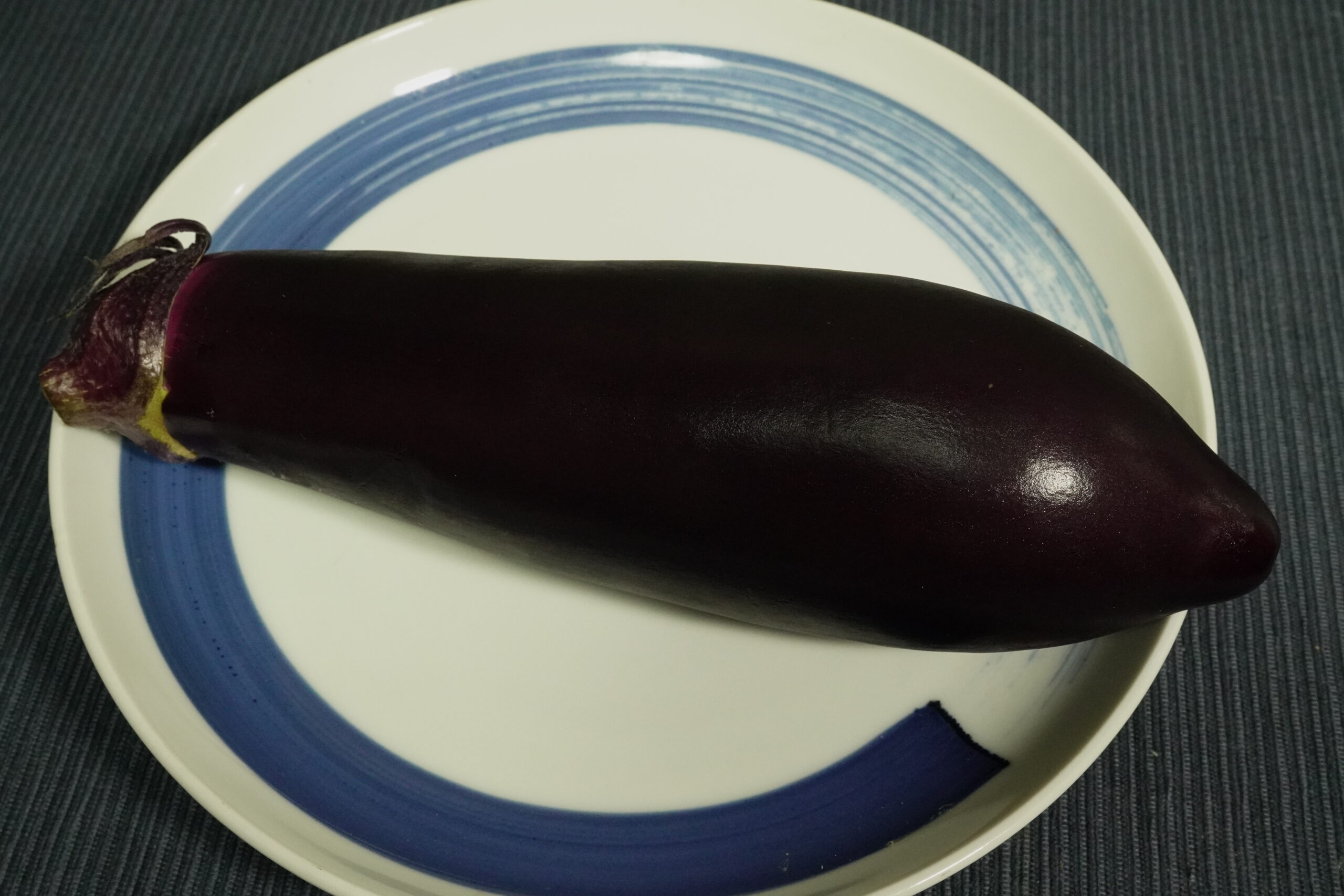 青と白の皿に置いた、皮が濃い紫色のなす1本の提供写真